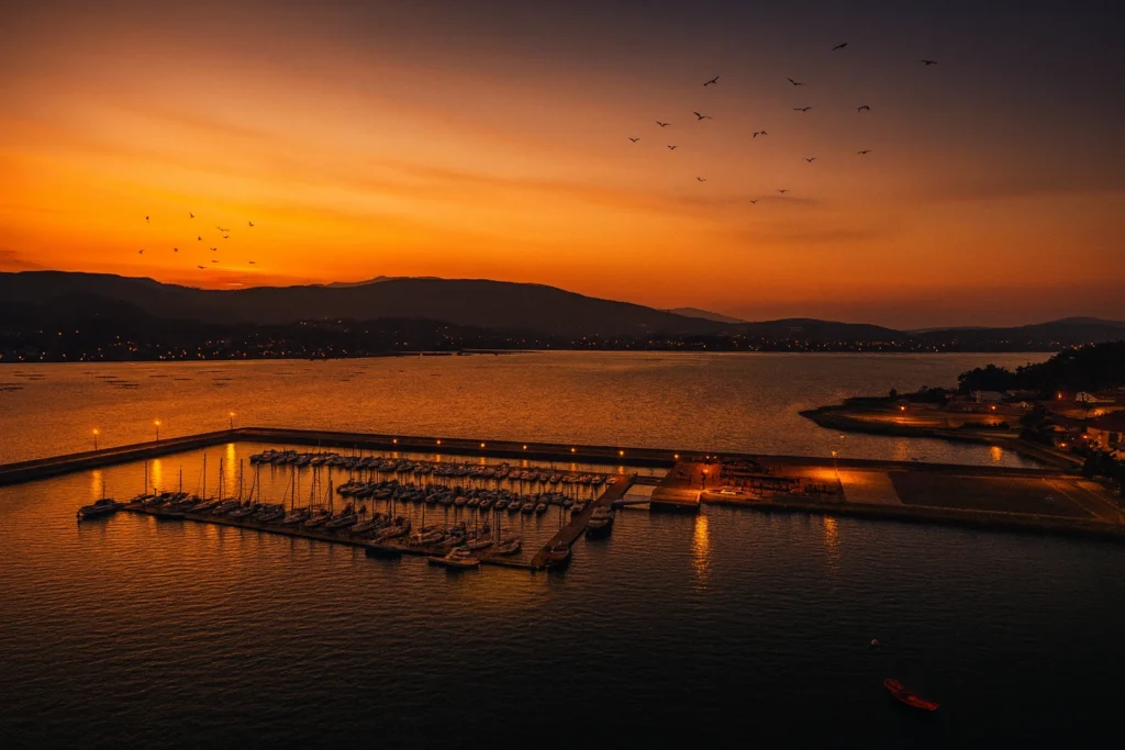 Vista aérea del Puerto Náutico de Boiro al atardecer, con los pantalanes iluminados, embarcaciones atracadas y un cielo anaranjado lleno de aves volando sobre la ría.